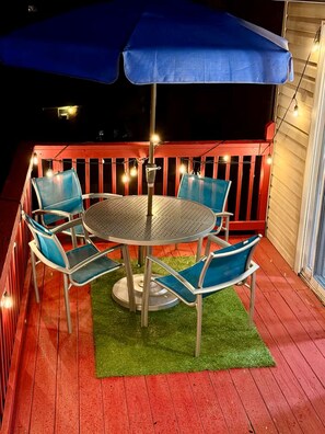 Een terras met een ronde tafel en stoelen, een blauwe parasol en lichtslingers.