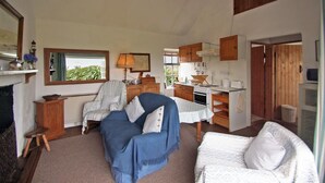 A cozy living room with a sofa, armchair, and a dining table. There is a kitchen area with cabinets and a stove. - 2 Bed in Ballyconneely (oc-i231) (Leitirshask)