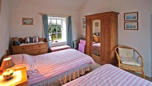 A bedroom with two beds, a wooden wardrobe, a wicker chair, a window with curtains, and a bedside table with a lamp. - 2 Bed in Ballyconneely (oc-i231) (Leitirshask)