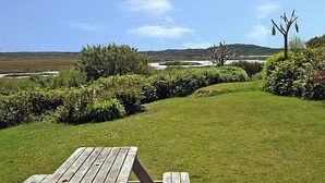 A grassy outdoor area with a wooden table and benches, surrounded by bushes and trees, with a view of hills and a body of water in the distance. - 2 Bed in Ballyconneely (oc-i231) (Leitirshask)