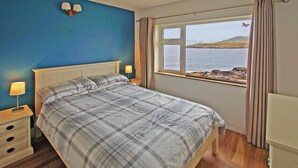 Un dormitorio con cama, lámparas de noche, mesita de noche, ventana con vista al mar y cortinas.