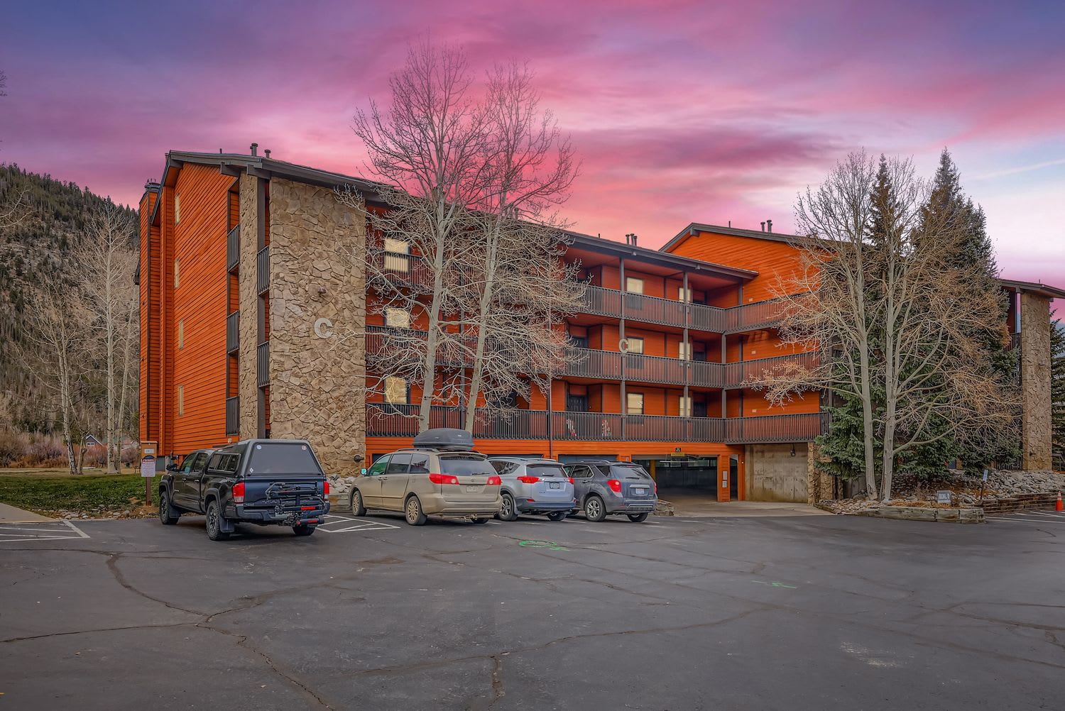 A multi-story building with a stone facade, a parking lot with several cars, and a mountainous backdrop.