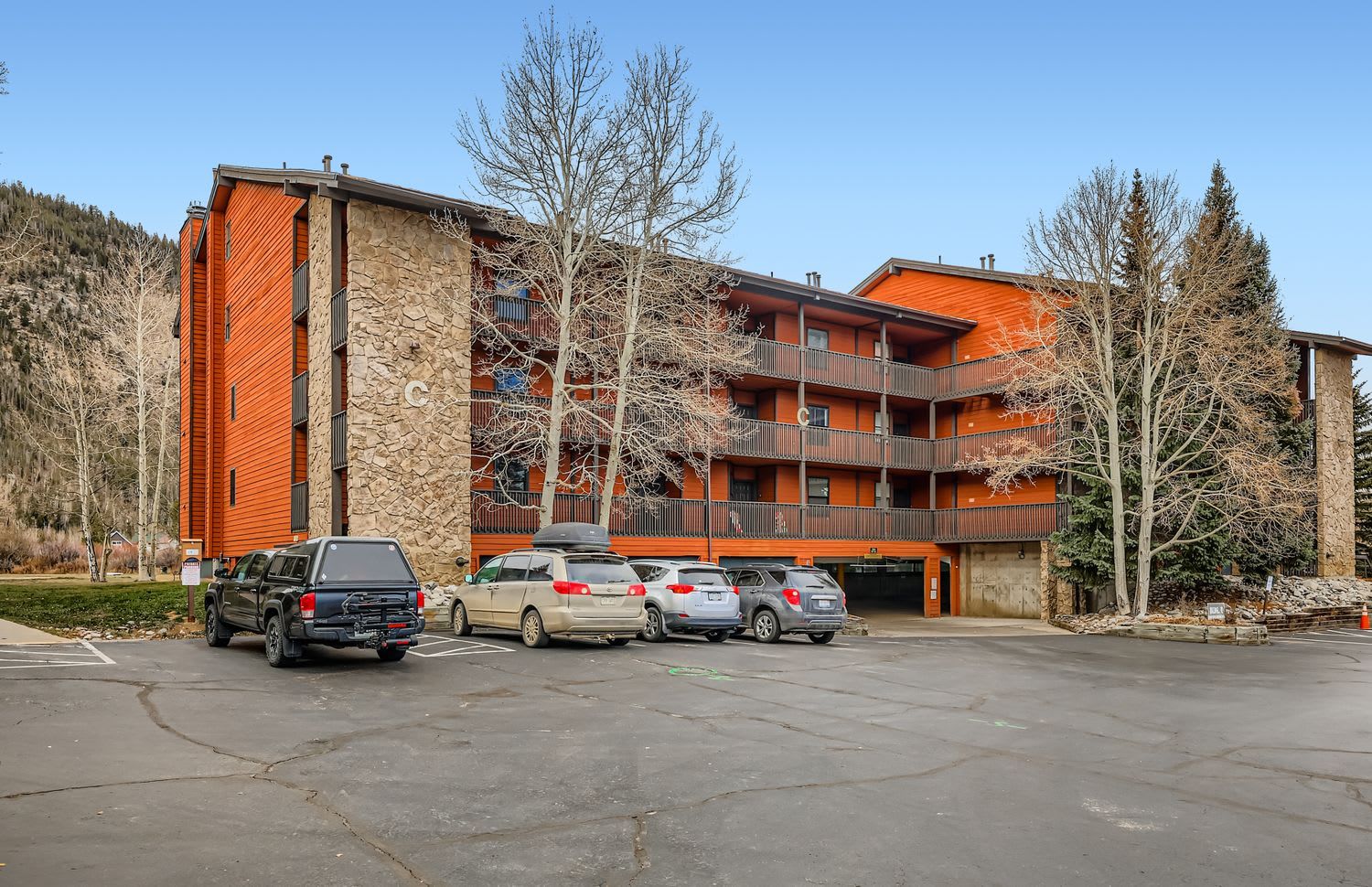 Un immeuble à plusieurs étages, doté d’une façade en pierre et de balcons, un parking avec des voitures et des arbres en arrière-plan.