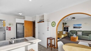 A modern kitchen with a built-in refrigerator, a dining area with a wooden table and chairs, and a living room with a sofa and a coffee table. - Get Away By The Green - Cromwell Holiday Home (Cromwell)