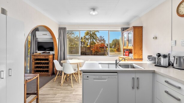 A modern kitchen with a dishwasher, a dining area with a table and chairs, and a large window with a view of trees. - Get Away By The Green - Cromwell Holiday Home (Cromwell)