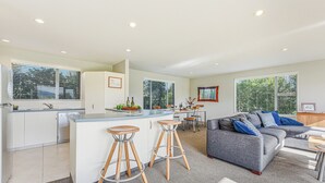 A modern kitchen with a central island, bar stools, a dining area, and a grey sofa with blue cushions. - Vineyard View - Blenheim Holiday Home (Blenheim)
