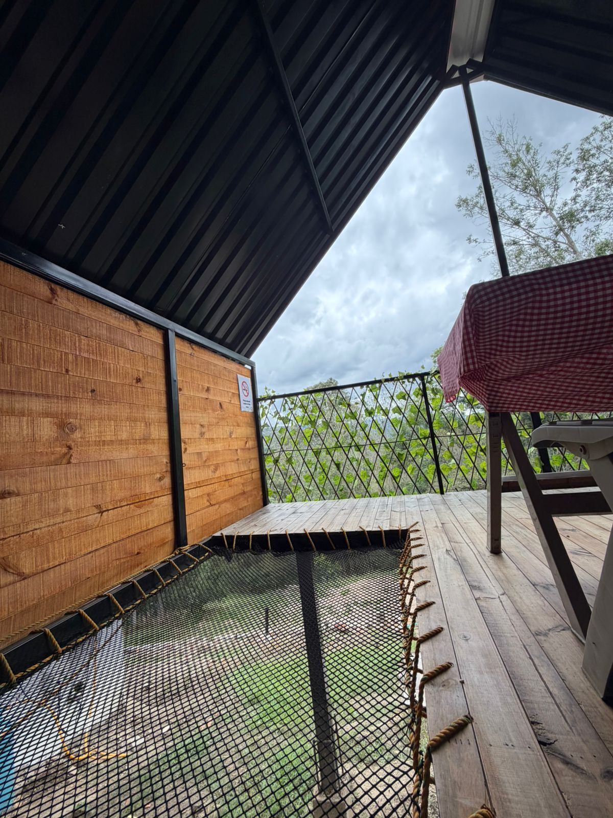 Un espace extérieur abrité, doté d’une terrasse en bois, d’un trampoline à mailles métalliques et d’une nappe à carreaux.