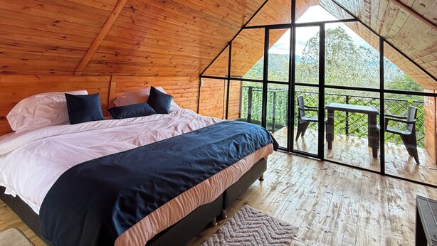 A wooden interior with a bed, a view through a glass wall, and a balcony with chairs.