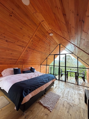 Un interior de madera con una cama, vistas a través de un muro de cristal y un balcón con sillas.