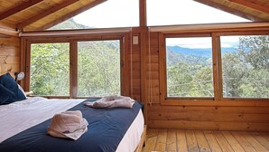 A wooden cabin bedroom with a bed, a nightstand, a lamp, and large windows offering a view of the outdoors.