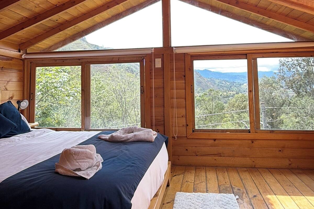 A wooden cabin bedroom with a bed, a nightstand, a lamp, and large windows offering a view of the outdoors.