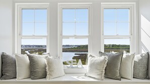 En sittplats vid fönstret med flera kuddar och en skylt med texten ”HAV”, med utsikt över en vattenyta och grönområden.