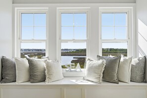 Sitzplatz am Fenster mit mehreren Kissen und einem Schild mit der Aufschrift 'SEA', mit Blick auf Wasser und Grünflächen.