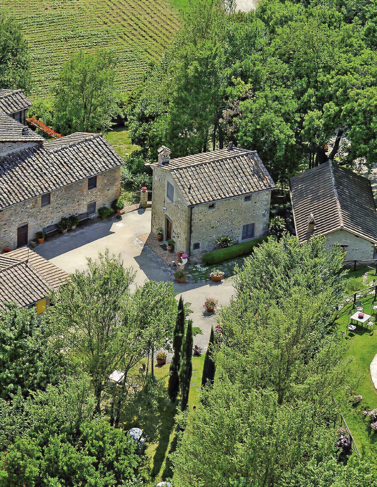 A stone building with a tiled roof surrounded by lush greenery and a vineyard in the background.