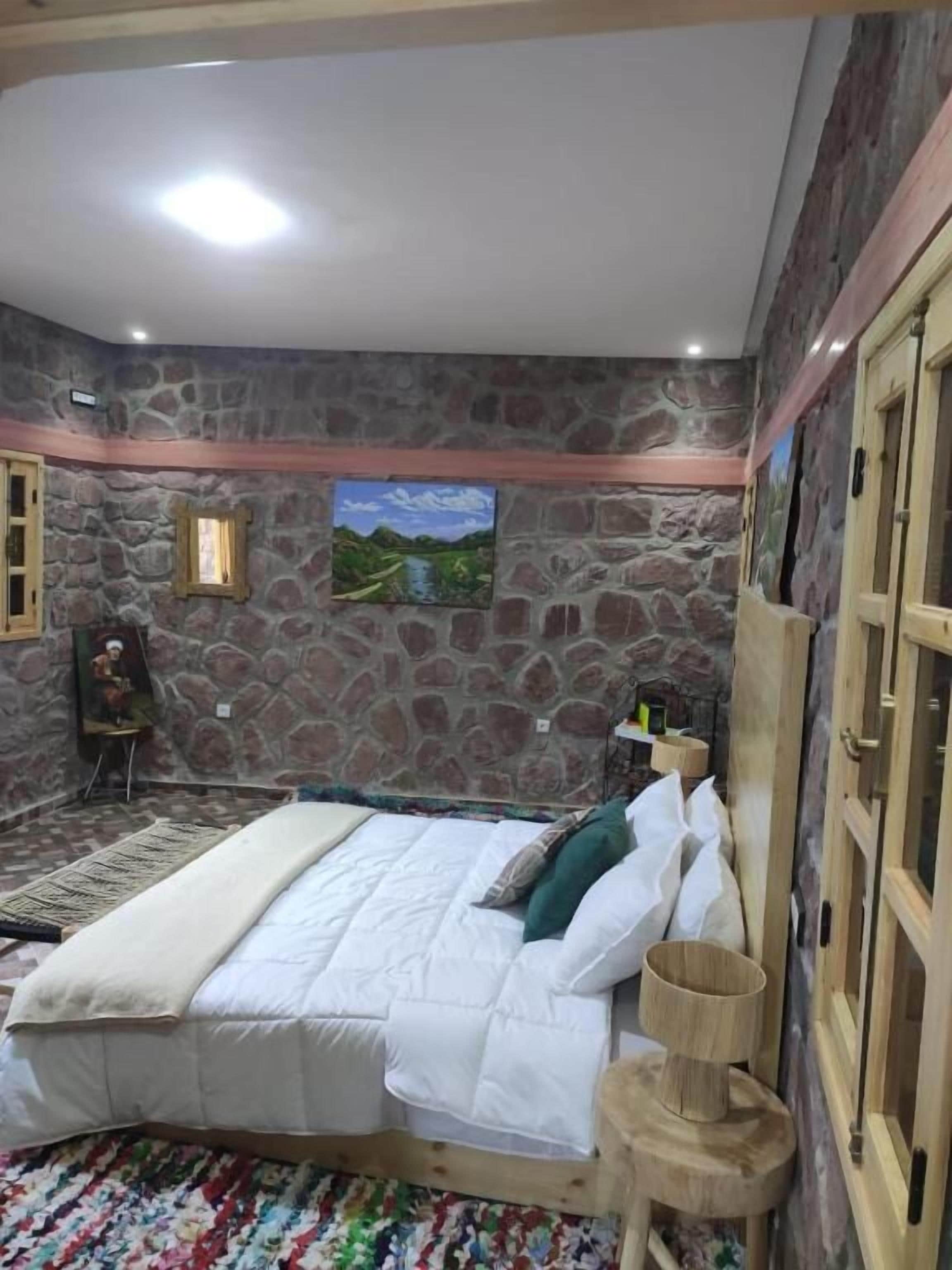 Un dormitorio con pared de piedra, una cama con ropa blanca, un cabecero de madera y una pequeña mesita de noche de madera.