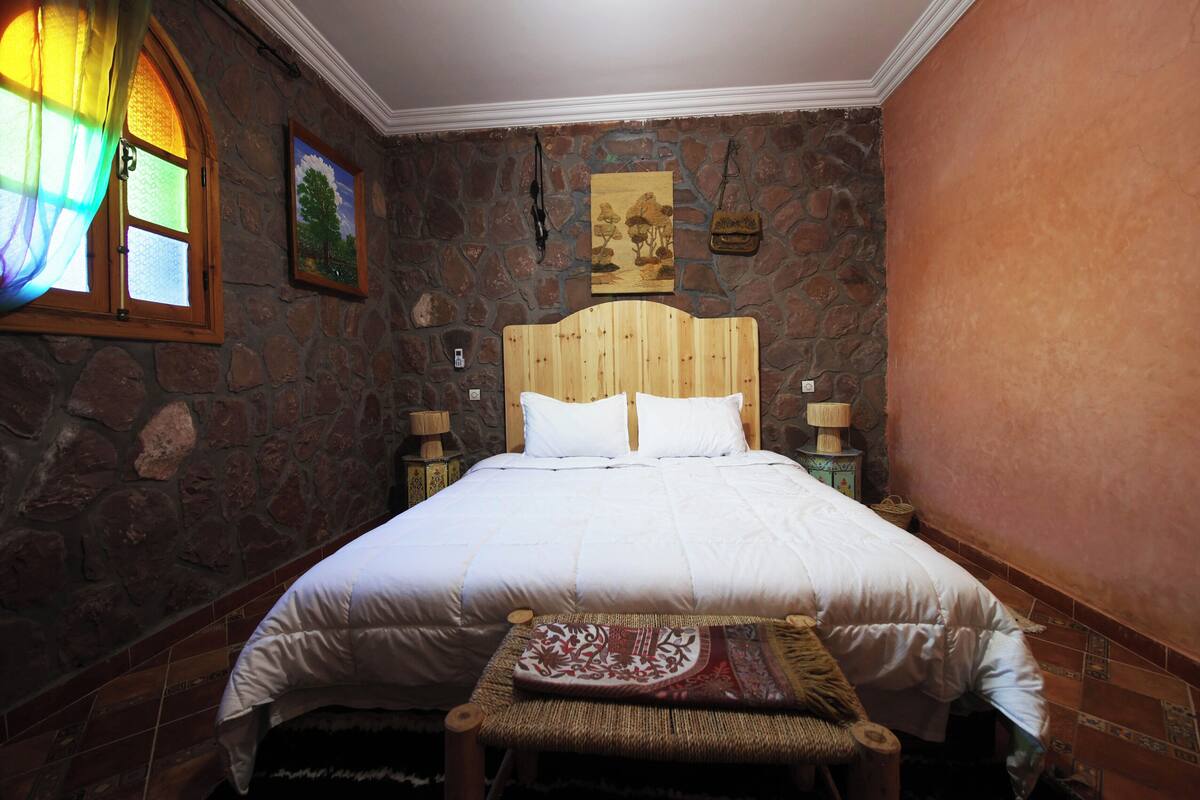 Un dormitorio con pared de piedra, cabecero de madera, cama con ropa blanca, mesita de noche con lámpara y un pequeño banco con cojín.