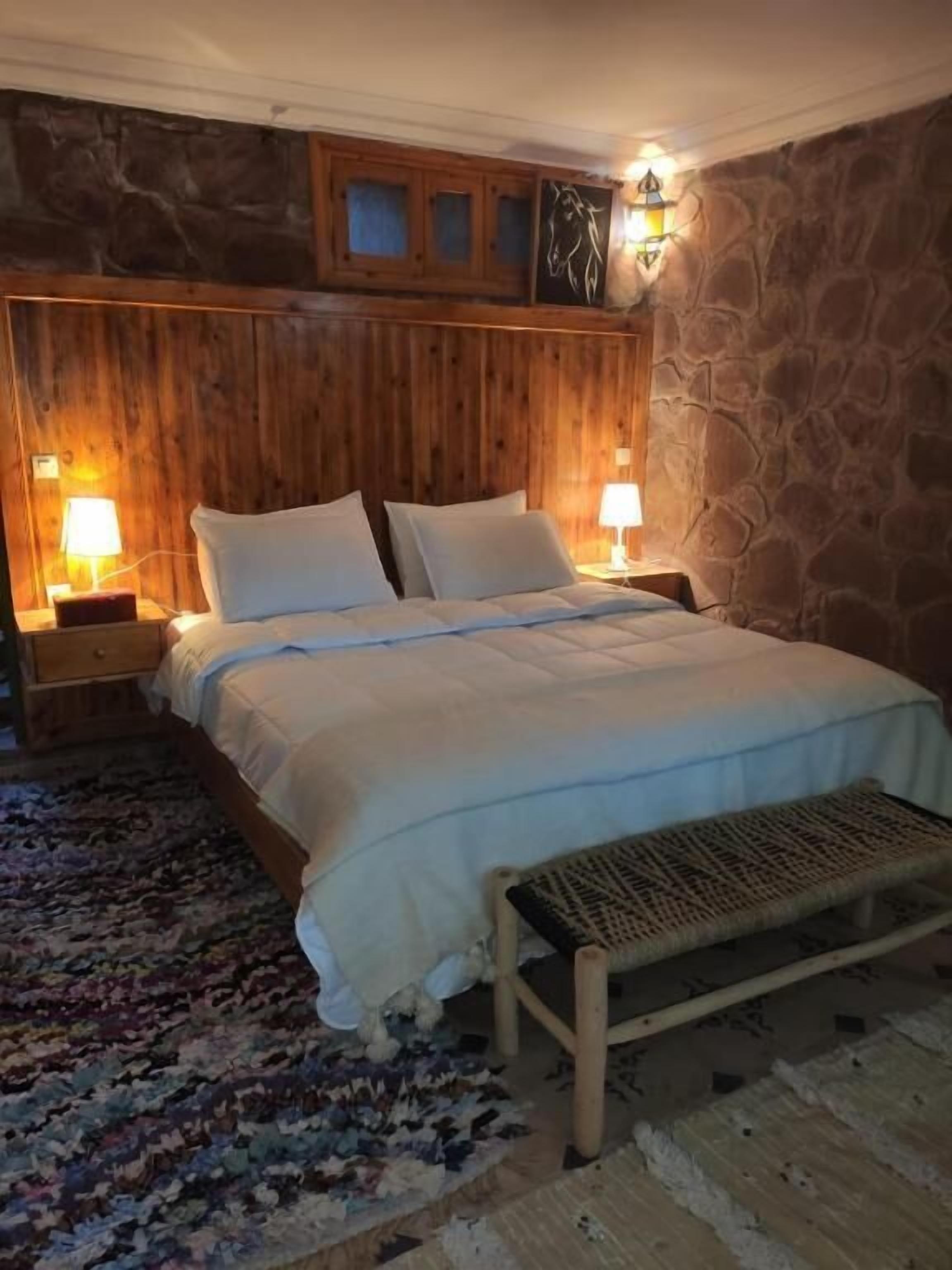 A bedroom with a wooden headboard, a bed with white linens, two bedside lamps, a stone wall, and a patterned rug.
