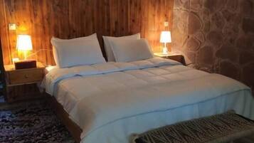 A bedroom with a wooden headboard, a bed with white linens, two bedside lamps, a stone wall, and a patterned rug.