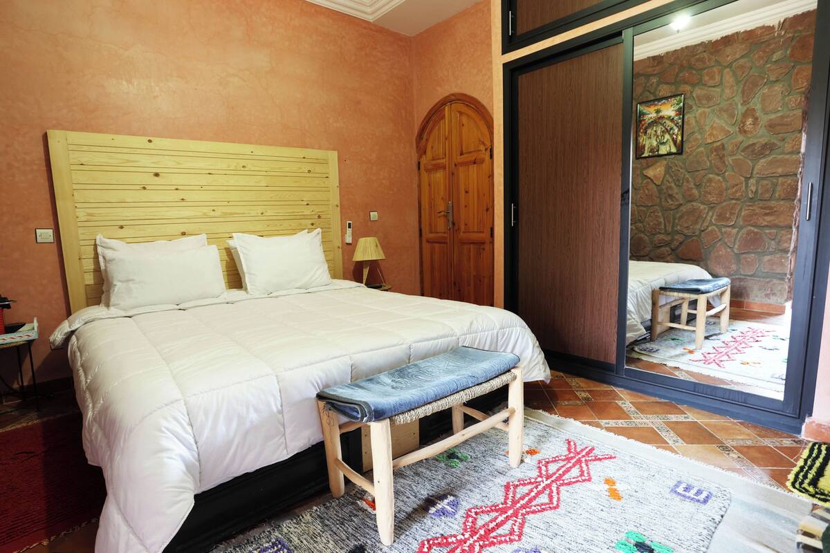 Un dormitorio con una cama, un banco de madera, una mesita de noche, una lámpara y una pared de piedra.