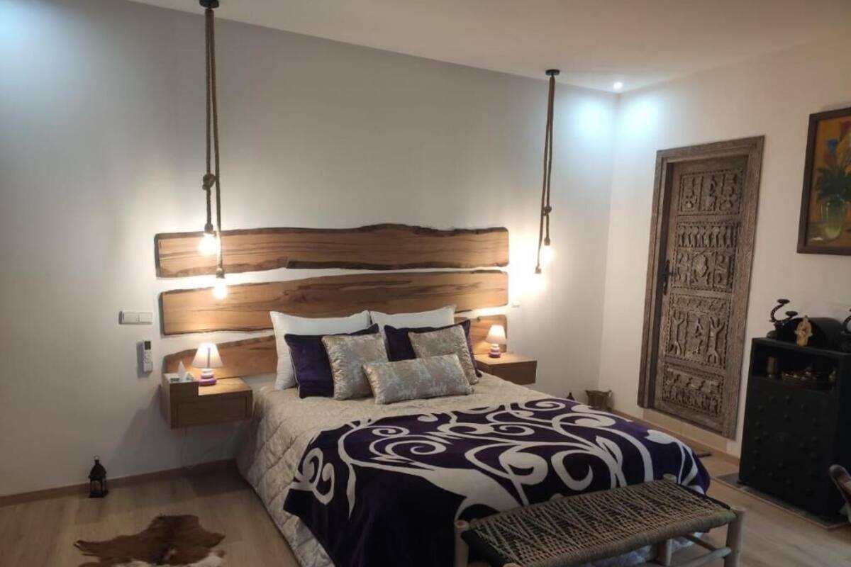 Un dormitorio con cabecera de madera, colcha estampada, lámparas de noche, un banco y un panel decorativo en la pared.