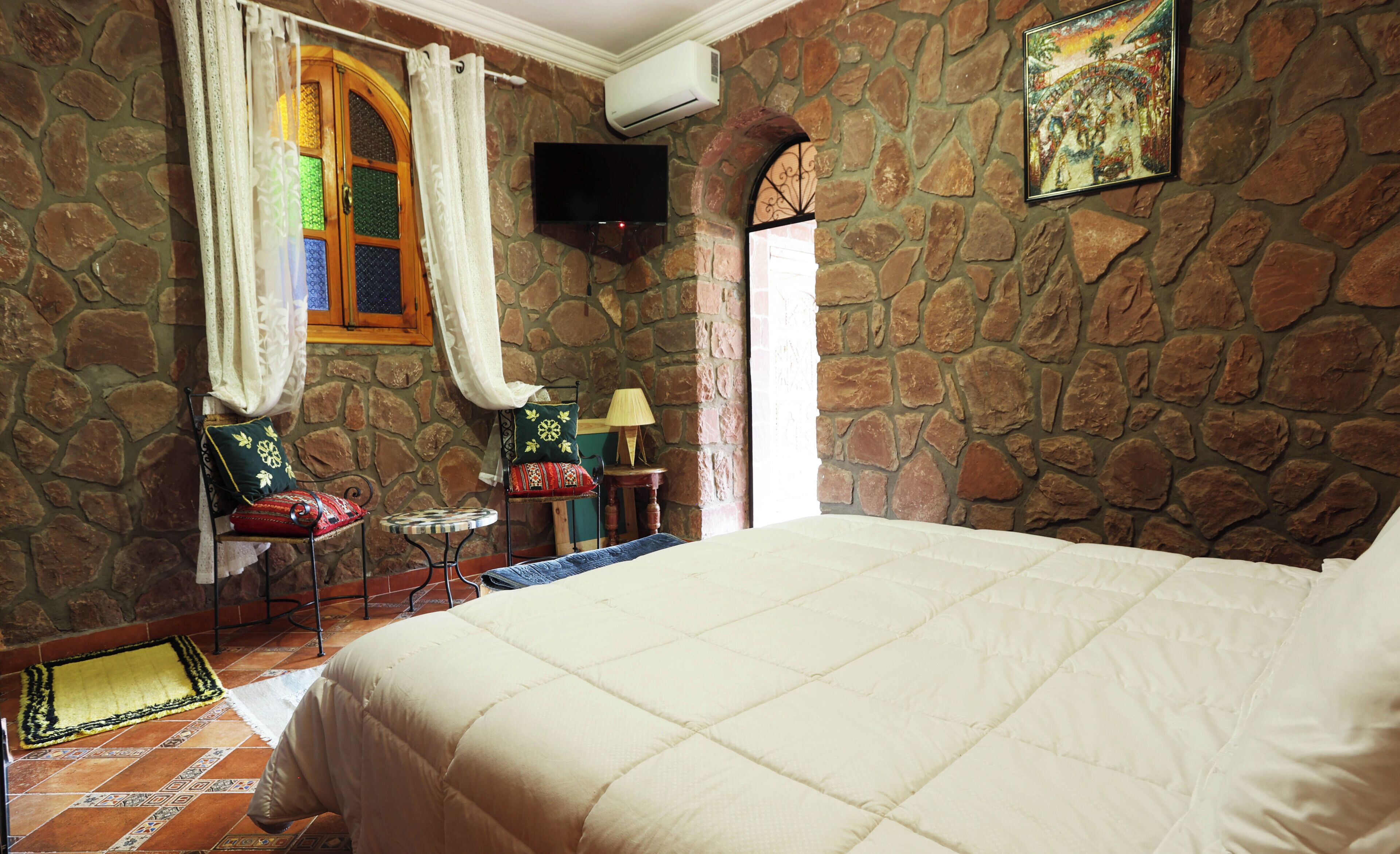 A room with a stone wall, a bed, a window with curtains, a small table with a lamp, and a painting on the wall.