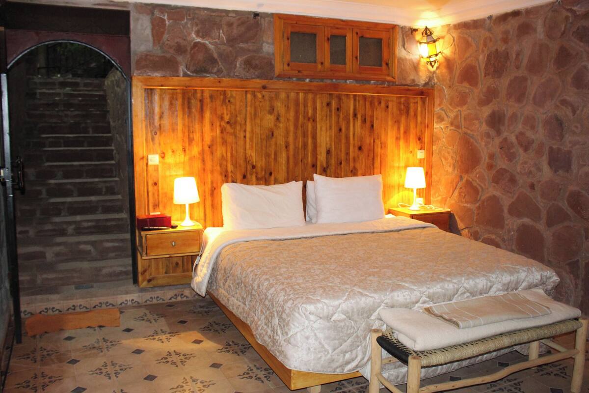 Un dormitorio con cabecera de madera, cama con sábanas blancas, dos lámparas de noche, un banco de madera y una pared de piedra.