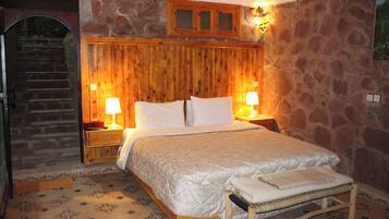 A bedroom with a wooden headboard, a bed with white linens, two bedside lamps, a wooden bench, and a stone wall.