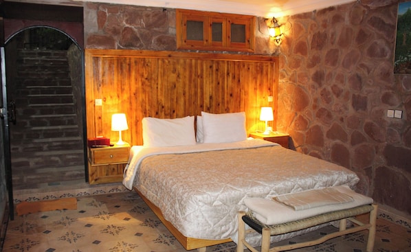 A bedroom with a wooden headboard, a bed with white linens, two bedside lamps, a wooden bench, and a stone wall. - Laurier rose (Sti Fadma)