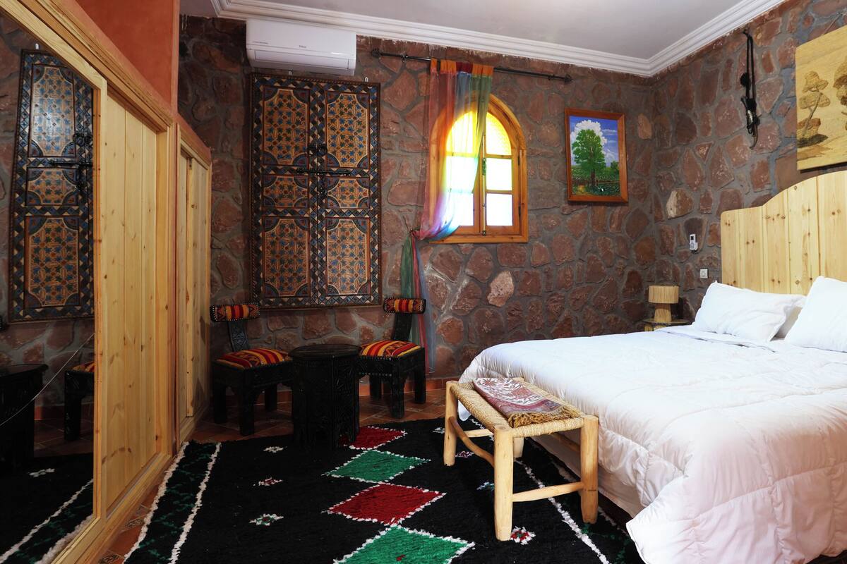 Una habitación con una cama, un banco, una mesita, una ventana con cortinas y una pared de piedra.