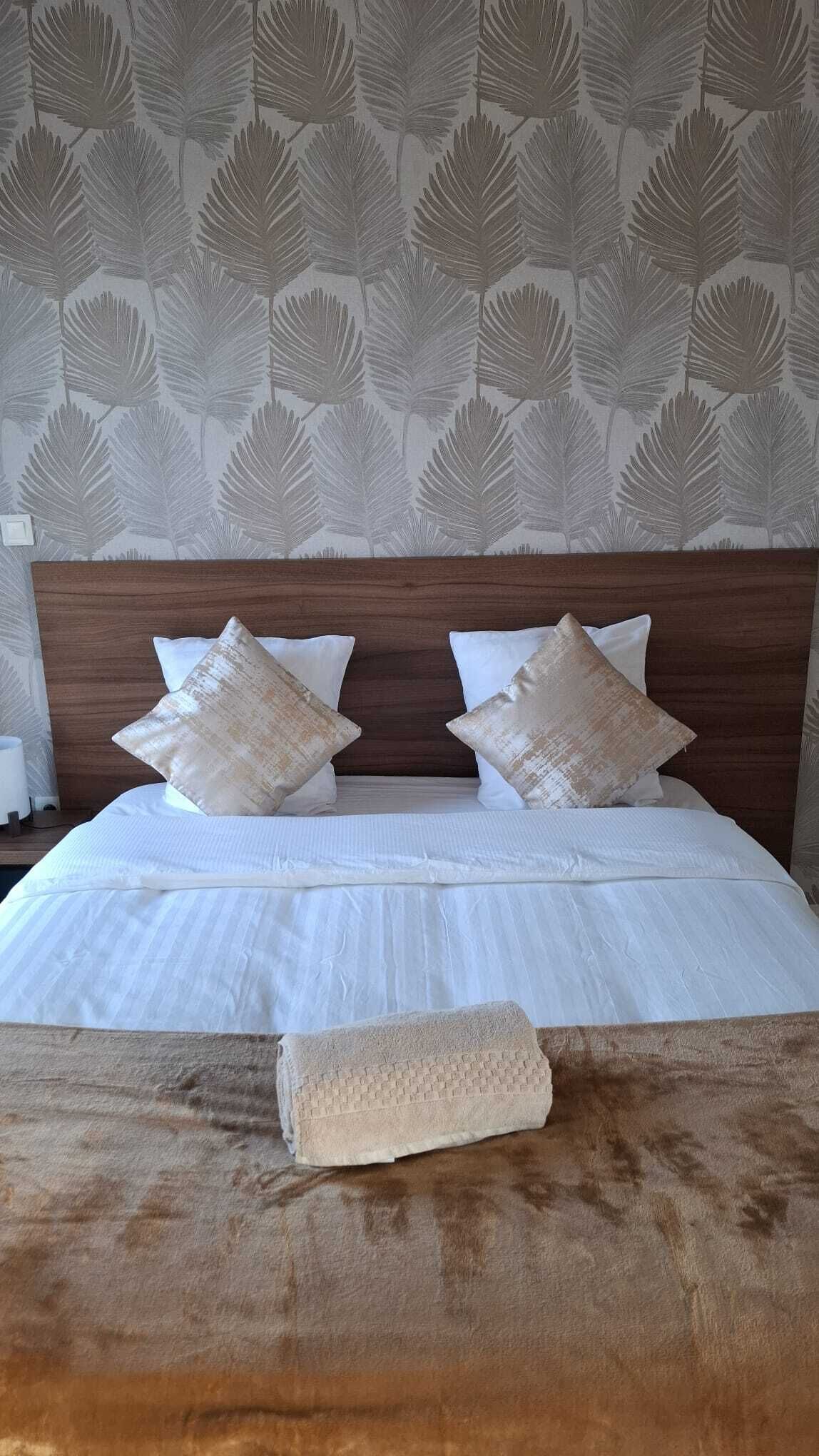 Una cama bien tendida con sábanas blancas y dos almohadones decorativos, un cabecero de madera y un papel tapiz con diseños de hojas.