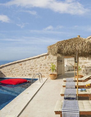 A poolside area with a stone wall, a thatched roof structure, lounge chairs, and a red cushion. - Villa Amarant - Five Bedroom Villa, Sleeps 10 (Zavala)