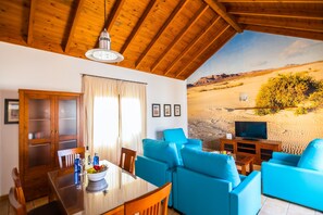 A room with a wooden ceiling, a dining table with chairs, a blue sofa, a TV, and a desert landscape mural on the wall. - Villa Lajares Dos - Two Bedroom Villa, Sleeps 4 (Lajares)
