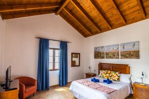 A bedroom with a wooden ceiling, a bed with a white bedspread, a chair, a desk with a computer, and a window with blue curtains. - Villa Lajares Dos - Two Bedroom Villa, Sleeps 4 (Lajares)