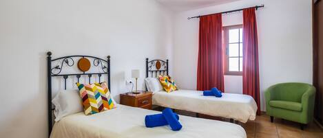 A hotel room with two beds, a green armchair, and a window with red curtains.