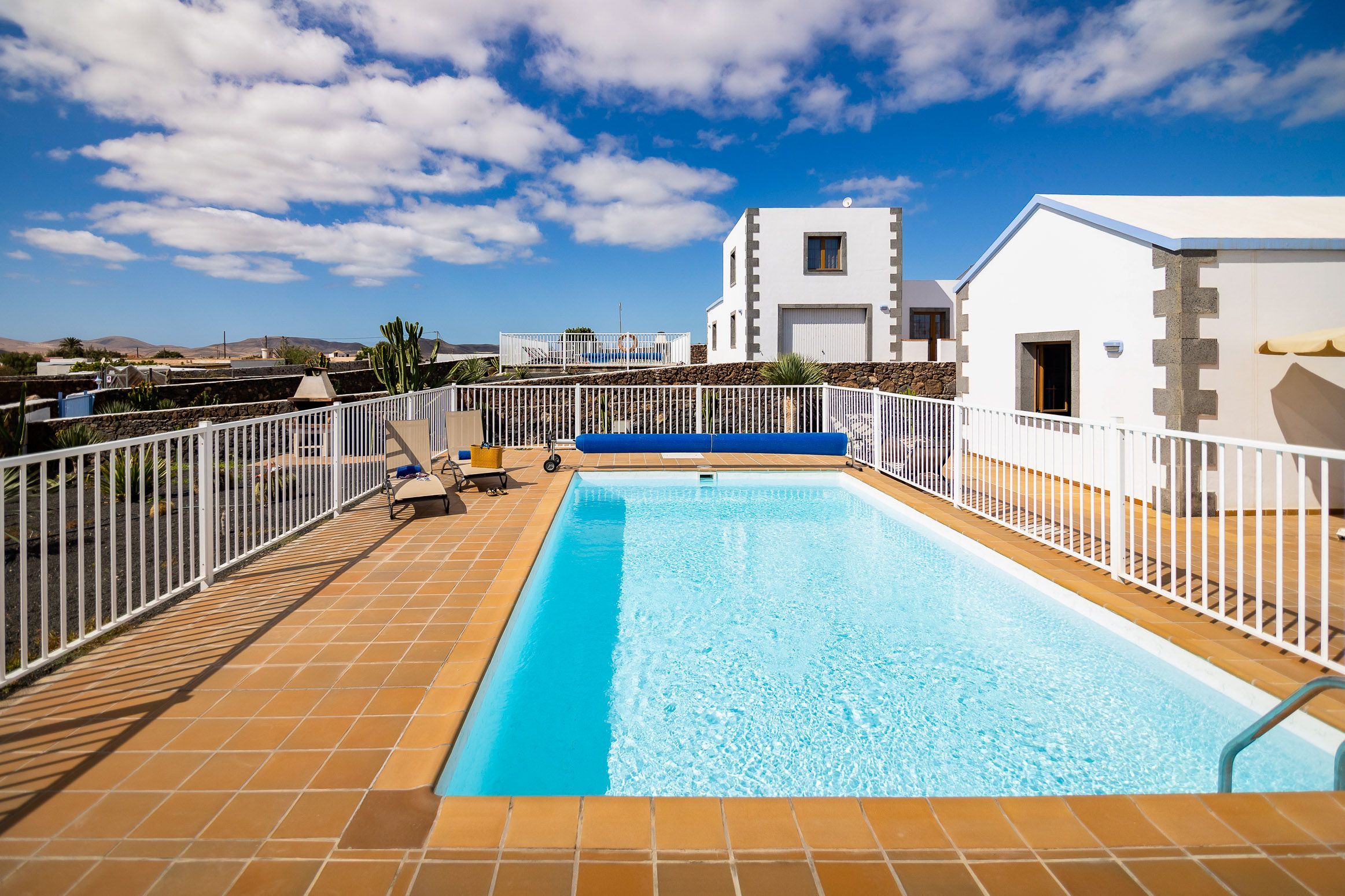 Piscina rectangular con una cerca blanca, frente a un cielo azul despejado y colinas lejanas.