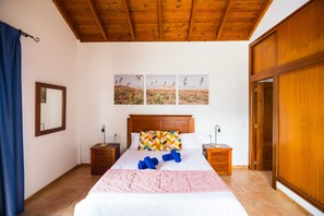 A bedroom with a wooden ceiling, a bed with a pink blanket, two bedside tables with lamps, and a wall with three framed pictures. - Villa Lajares Tres - Three Bedroom Villa, Sleeps 6 (Lajares)