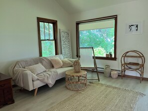 A cozy room with a sofa, a small round table, a rattan chair, a large window with a view of trees, and a framed picture on the wall.