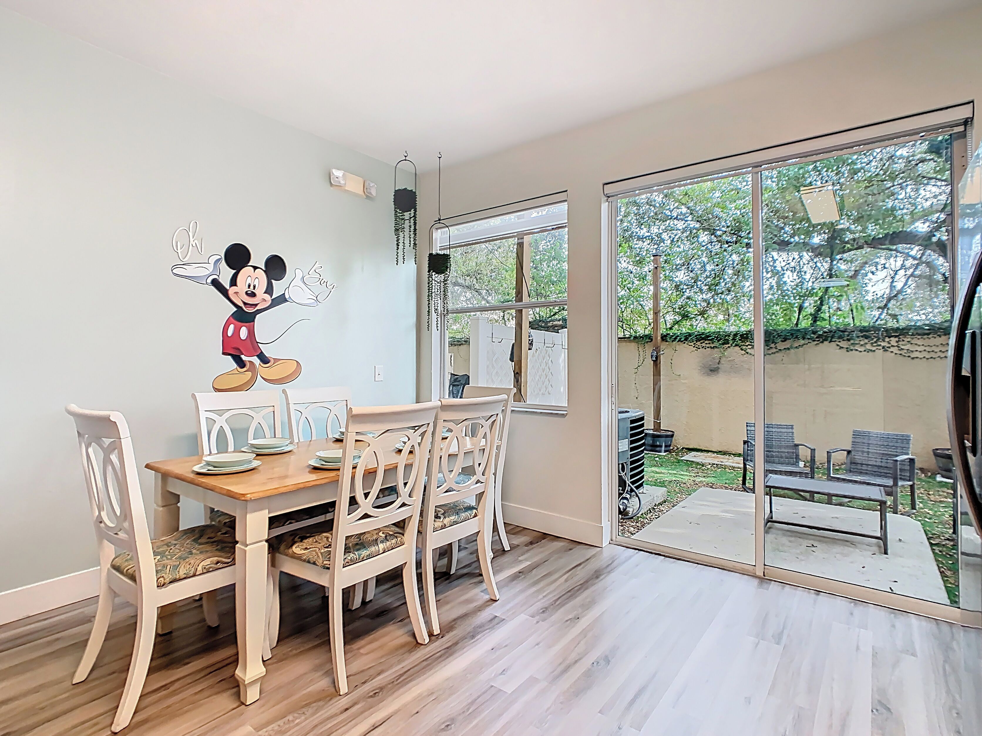 Un coin repas avec une table et des chaises en bois, un autocollant mural représentant Mickey Mouse, et une porte-fenêtre coulissante donnant sur un patio extérieur aménagé avec des sièges.