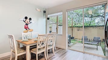 Un coin repas avec une table et des chaises en bois, un autocollant mural représentant Mickey Mouse, et une porte-fenêtre coulissante donnant sur un patio extérieur aménagé avec des sièges.