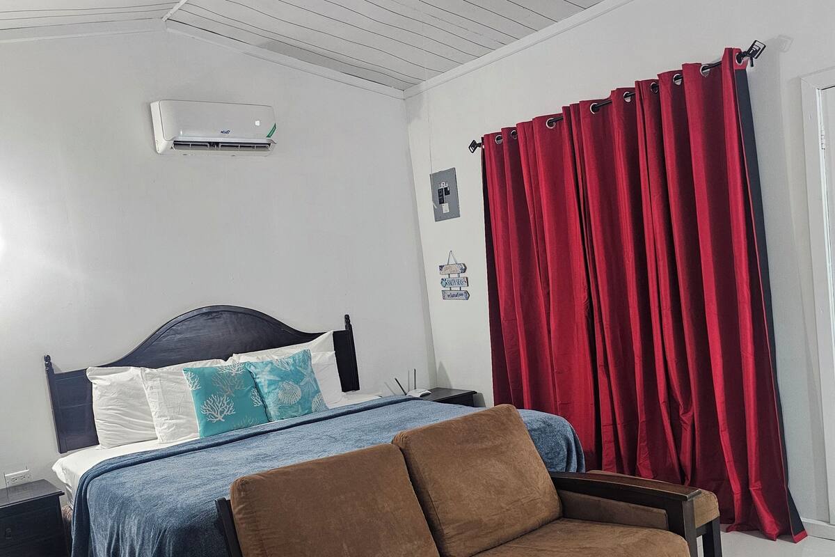 A bedroom with a bed, a sofa, a ceiling fan, a red curtain, and a wall-mounted air conditioner.