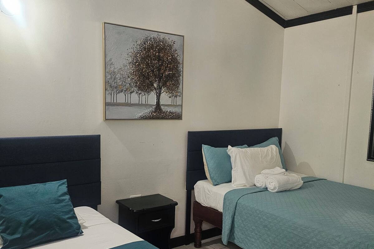 A hotel room with two beds, a nightstand, a ceiling fan, and a painting of a tree.