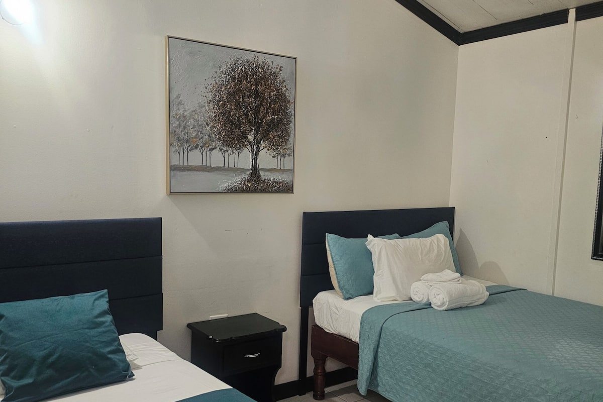 A hotel room with two beds, a nightstand, a ceiling fan, and a painting of a tree.