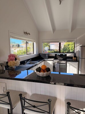 A modern kitchen with a central island, white cabinetry, and a view of the outdoors. - Hosts on the Coast Crown Reef Cottage (Pauanui)
