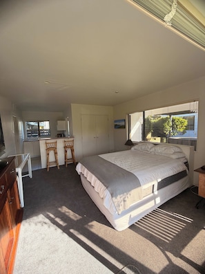 A bedroom with a bed, a desk, and a chair. There is a window with blinds and a view of trees outside. - Hosts on the Coast Crown Reef Cottage (Pauanui)