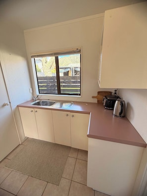 A small kitchenette with a window, a sink, cabinets, a toaster, and a kettle. - Hosts on the Coast Crown Reef Cottage (Pauanui)