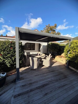 A covered outdoor patio with a round table and wicker chairs, surrounded by greenery and a wooden deck. - Hosts on the Coast Crown Reef Cottage (Pauanui)