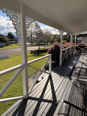 A covered outdoor area with a wooden deck, white railings, and a view of a grassy area with trees and a road. - Hosts on the Coast Crown Reef Cottage (Pauanui)