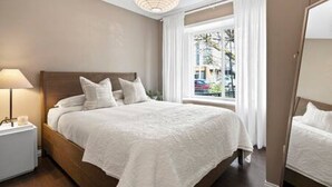 A neatly made bed with white linens, a bedside lamp, and a window with curtains. - Chic 2BR Mount Pleasant Suite Near Downtown (Vancouver)