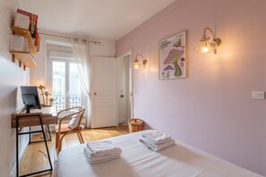 Ein Hotelzimmer mit einem Bett, einem Schreibtisch mit Computer, einem Stuhl, einem kleinen Tisch, einer Lampe und einem Poster an der Wand.