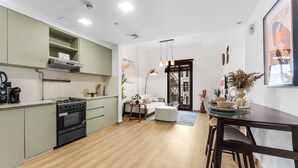 A modern kitchen with a stove, oven, and green cabinets. There is a dining area with a table and chairs, and a sofa. - Free Housekeeping Visits During Your Stay - Stayshort - Cozy Townsquare 1 Bed Apartment With Balcony That Sleeps 4 (Dubai)